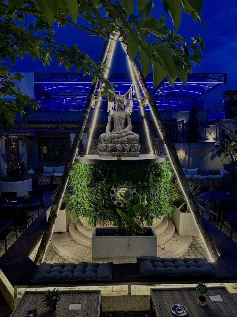 Il giardino del Blanco Garden illuminato di sera, con una statua di Buddha su una parete di verde e divanetti attorno