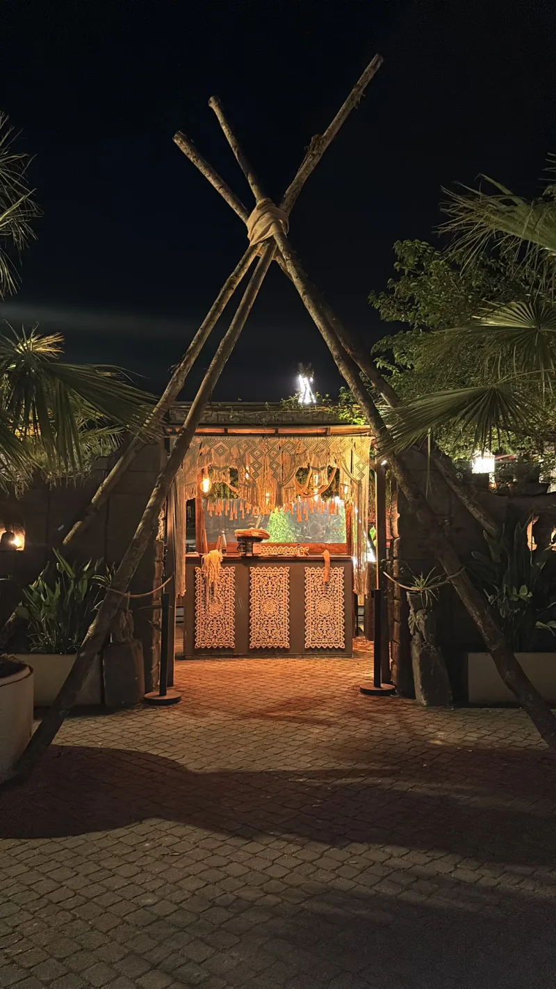 L'ingresso del Blanco Garden di sera: un bar in legno intagliato sotto una struttura di pali incrociati, tra palme e lucine